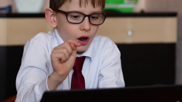 Attractive schoolboy 7 years old in glasses is studying online while at home in front of a gadget.