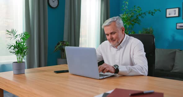 Serious Senior Man Sit at Cozy Home Office Table Working on Laptop 55s Businessman Consider