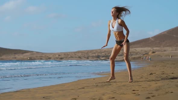 Girl in Sportswear Shorts and T-shirt Performs Jumps with Squats on the Beach Near the Ocean in the