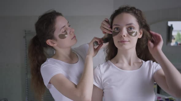 Confident Beautiful Woman with Eye Mask Looking at Camera As Twin Sister Brushing Hair of Sibling