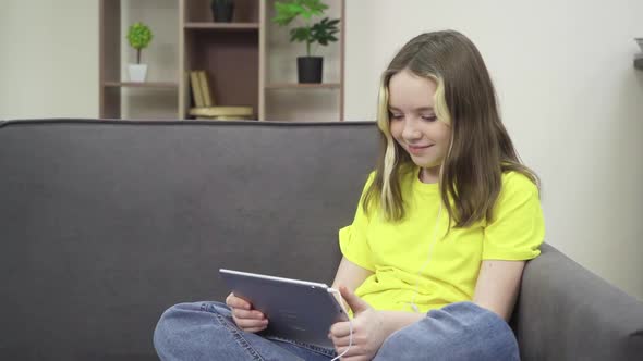 The Little One Smiles and Looks at the Tablet While Sitting on the Couch