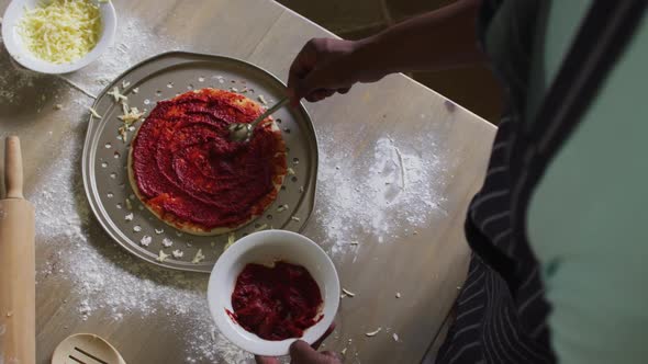 African american girl making pizza spreading tomato on base in kitchen