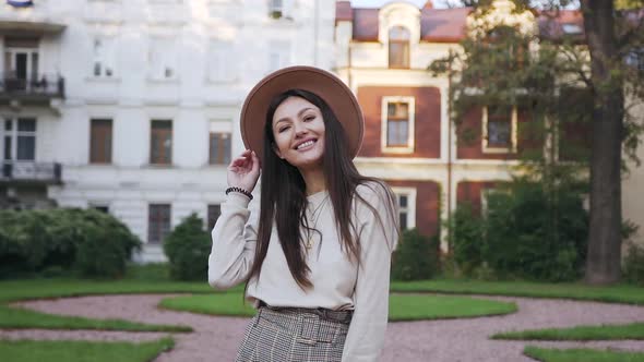 Woman in Trendy Hat Posing on Camera with Sincerely Smile in Front of Beautiful Mansions