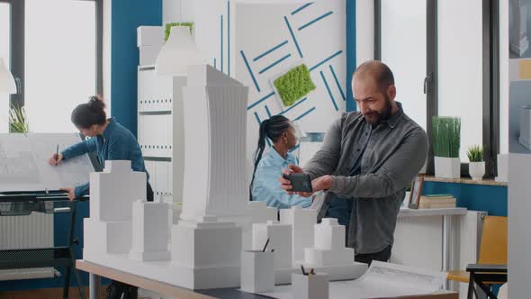 Man Engineer Using Smartphone to Work on Building Model and Layout in Architectural Office