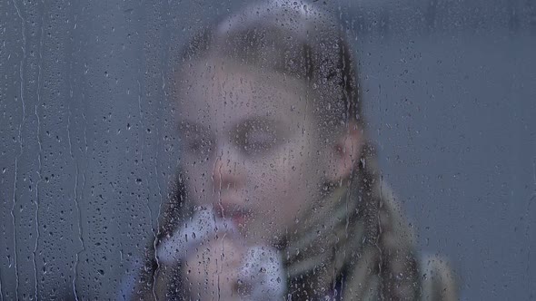 Little Girl Sneezing and Looking to Camera Through Rainy Window