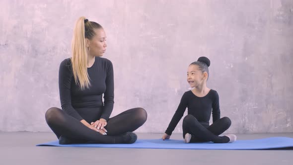 Beautiful Young Gymnastics Teacher and Little Asian Girl Sitting on Yoga Mat in Studio and Talking