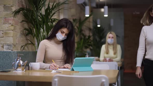 Focused Slim Beautiful Middle Eastern Woman in Covid Face Mask Sitting at Table with Tablet Writing