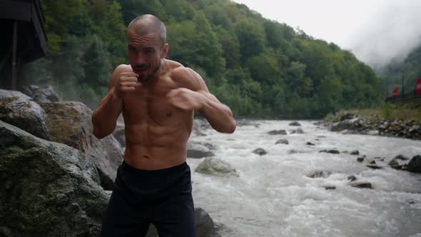 an Unshaven Man with a Bare Torso is Boxing By the River Against the Background of Mountains