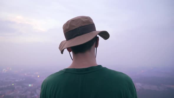 The camera films a man from behind. The model looks at the city of Nanjing, China. He wears a bucket