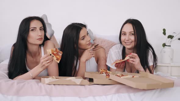 Three Gorgeous Girlfriends Are Lying on the Bed and Eating Delivery Pizza