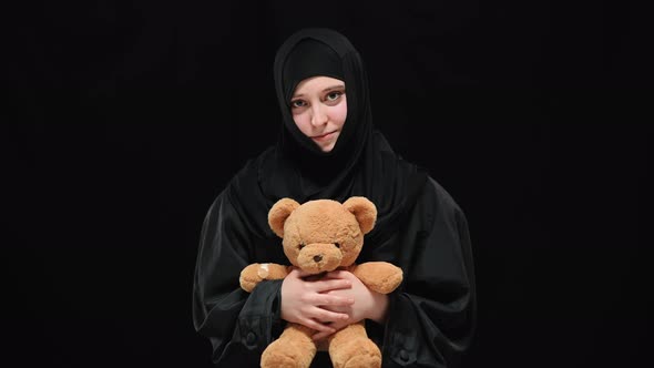 Middle Shot Portrait of Shy Muslim Woman in Hijab Posing at Black Background with Teddy Bear