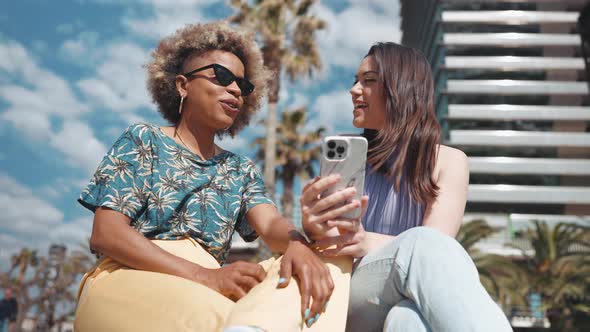 Smiling sunny multiethnic women talking and looking at phone outdoors