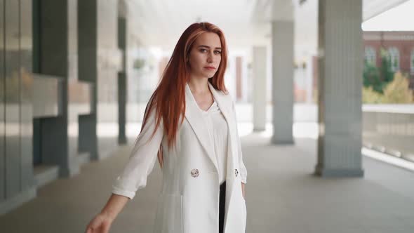 Beautiful business woman red hair in white coat stands near an office building