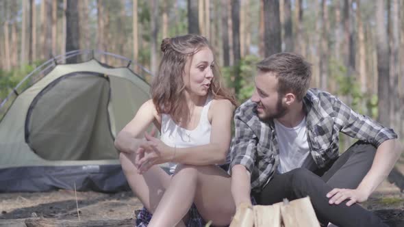Portrait Young Man Kindling a Fire in the Forest While Adorable Young Woman Sitting Near. Loving