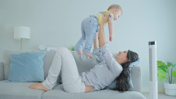 Happy Caucasian loving mom play with baby boy child on sofa in living room in morning at house.