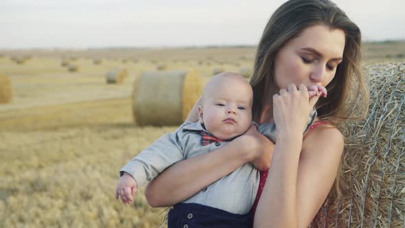 Happy Mother Kisses Baby's Little Hands When Holds Him on Hands in the Hay Field