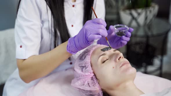 Beautician Applies a Black Mask on the Face of the Client's Face in a Beauty Salon