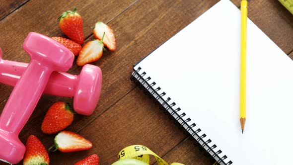 Strawberry, measuring tape, dumbbells and spiral notebook on table