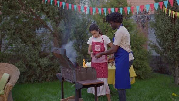 Wide Shot of Concentrated African American Teenage Daughter Helping Father Setting Fire on Barbecue