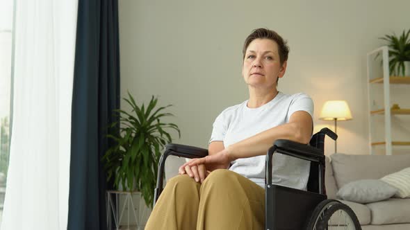 Portrait of Mature Sad Woman Sitting in Wheelchair at Home and Looking at Camera