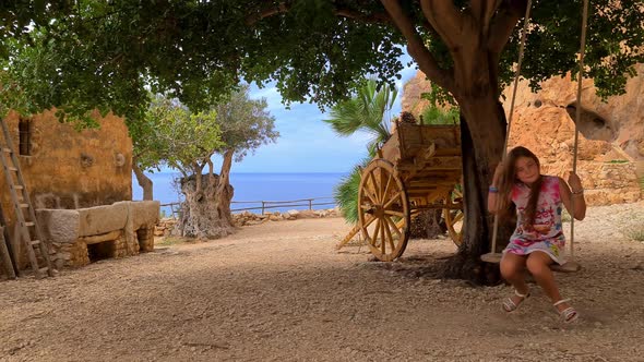 Cute redhead girl have fun swinging on swing tied to tree with Sicilian cart or carretto siciliano i