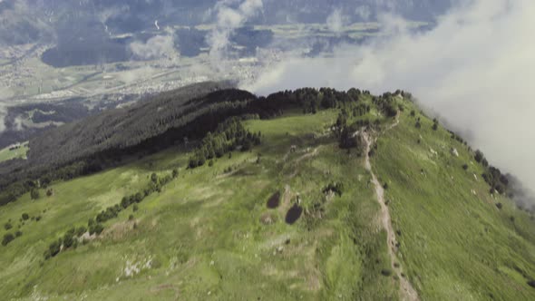 Aerial drone shot of a grass covered mountain top with a small trail leading up with clouds around.