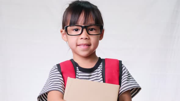 Smiling schoolgirl wearing summer outfit with backpack holding books on white background in studio.