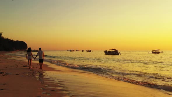 Couple in love walking around exotic beach washed by sea flows reflecting yellow sky of twilight ove