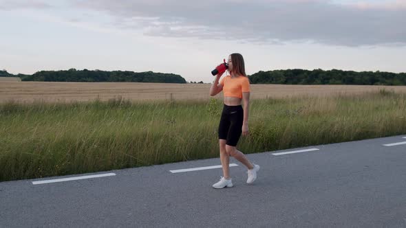 Athletic Girl Walking on Path at Nature in Morning and Drinking From Sport Flask