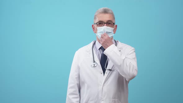 Man Doctor in Uniform with Glasses Removes a Medical Mask with Hand and Sighs Lightly in the Studio