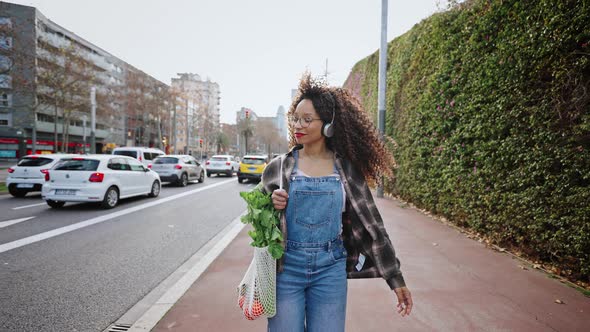 Lovely Student Walks Along a City Street at Dawn with a Bag of Oranges and Herbs and Listens to