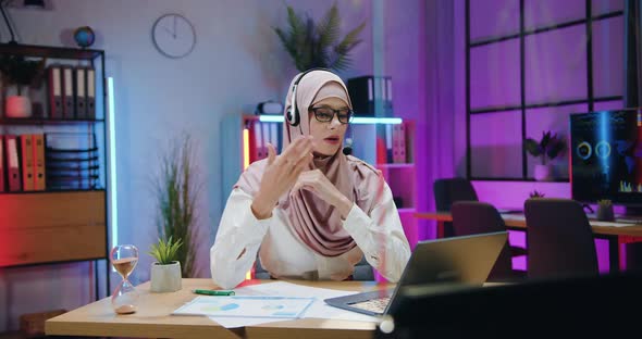 Arabian Businesswoman in Hijab Wears Headset During Online Conference on Laptop in the evening
