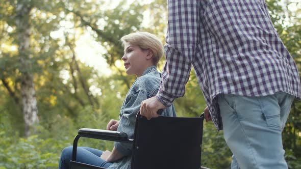 Man Rolls Emotional Disabled Woman in Wheelchair Through Park and Talks to Her Side View Closeup