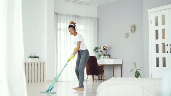 Cheerful African American Girl Is Mopping Floor, Listening To Music with Wireless Headphones and