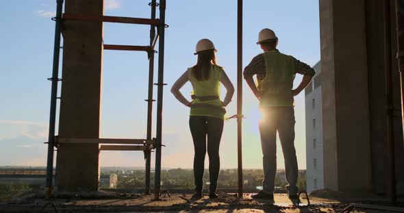 Builders Look at a Cityscape on a Sunset Background Back View