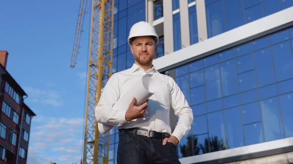 An Engineer in a White Shirt and Helmet Stands Against the Background of a Modern Glass Building and