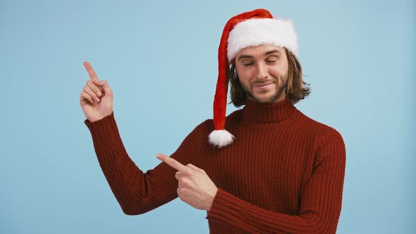 Young Male in Santa Hat is Pointing Up at Something Winking and Smiling While Posing Against Blue