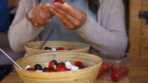 Caucasian woman cutting cherry tomatoes to a bamboo bowl plate with feta cheese and olives