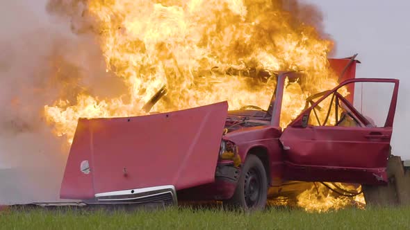 A red car explodes in an open field. The car is on fire.