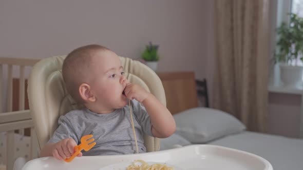 Toddler independently eats spaghetti with his hands while sitting on a feeding chair