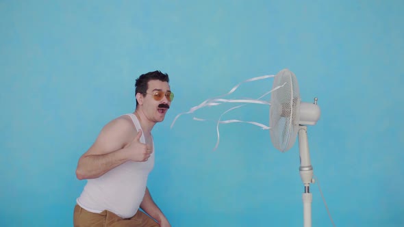 Funny Young Man with an Electric Fan Enjoying the Cool Breeze Showing Thumbs Up on Blue Background