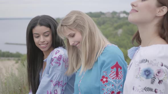 Three Girlfriends in Beautiful Long Summer Dresses with Embroidery Sitting and Talking on the River