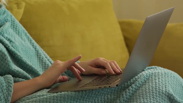 Close Up of a Caucasian Woman Hands Typing on a Laptop Computer Under Blue Blanket on a Yellow Sofa