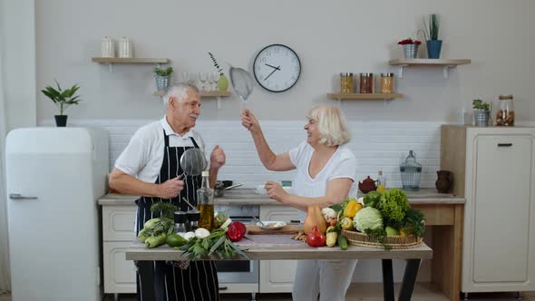 Senior Woman and Man Making a Funny Dance with Strainers. Dancing While Cooking Together in Kitchen