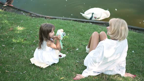 Talented Curios Little Girl Photographing Woman Sitting on Lawn on Lake Shore with White Swan