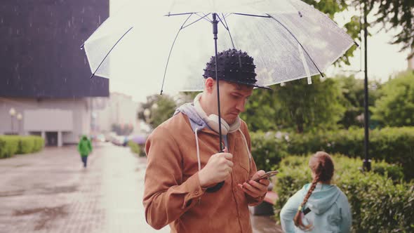 Young Handsome Man Browsing Mobile Phone Standing with Transparent Umbrella in Rain on City Street