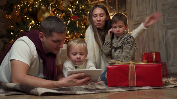 Happy Young Parents with Young Children are Sitting on the Warm Floor Near the Christmas Tree Making