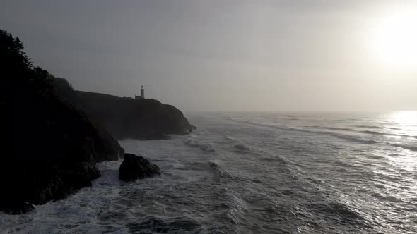 Huge waves burst on a rugged pacific ocean coastline, a silhouetted lighthouse in the distance, aeri