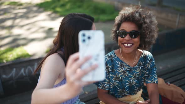 Two cheerful multiethnic women making selfie by phone while sitting on the bench