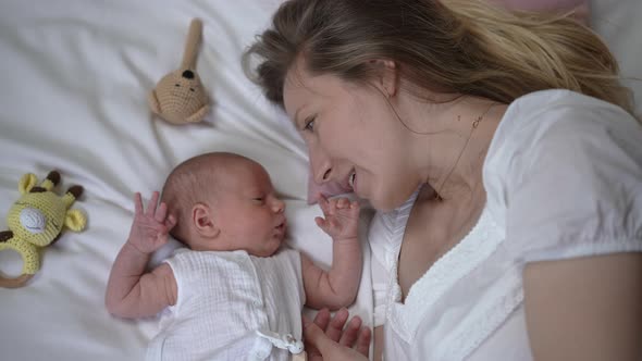 Happy Woman Talking with Newborn Boy Lying on Comfortable Soft White Bed at Home